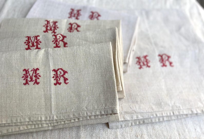 May include: Several off-white linen napkins with red embroidered initials "MR". The napkins are folded and stacked, with some visible in the background. The fabric has a textured appearance.