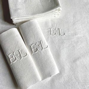 May include: White linen napkins and a tablecloth with an embroidered "EL" monogram. The napkins are stacked and folded, with two rolled up. The tablecloth and napkins have a subtle floral pattern.