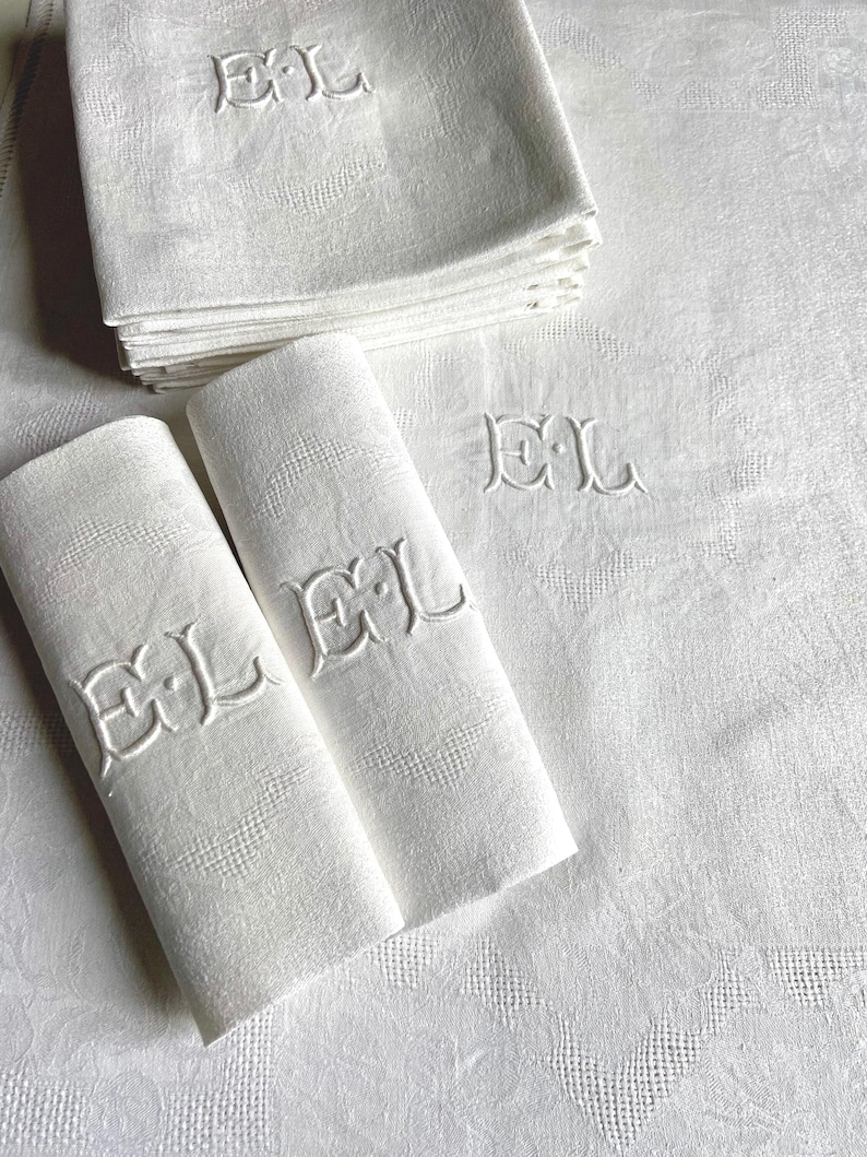 May include: White linen napkins and a tablecloth with an embroidered "EL" monogram. The napkins are stacked and folded, with two rolled up. The tablecloth and napkins have a subtle floral pattern.