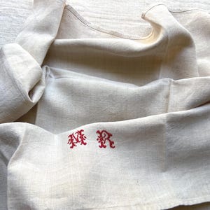 May include: Cream-colored linen textile with red embroidered initials. The fabric is folded and draped, showcasing its texture and the embroidered design. The initials are in a decorative, stylized font.