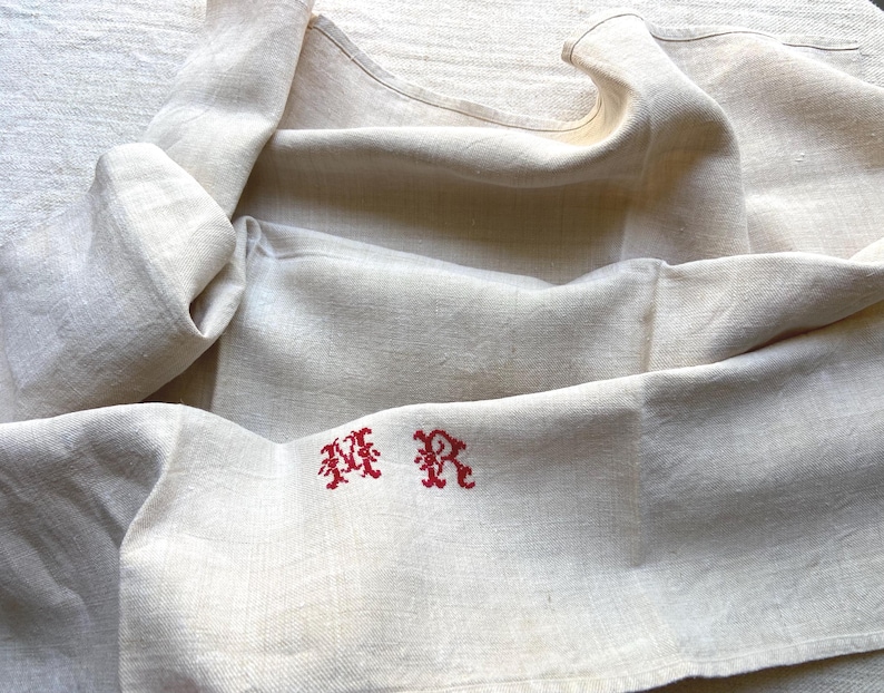 May include: Cream-colored linen textile with red embroidered initials. The fabric is folded and draped, showcasing its texture and the embroidered design. The initials are in a decorative, stylized font.