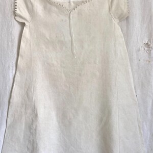 Puede incluir: Un vestido recto de color blanco roto con mangas cortas, que presenta un escote festoneado con un borde delicado. La prenda tiene una silueta sencilla y recta con costuras verticales en la parte delantera y un fino lazo en el centro del cuello.