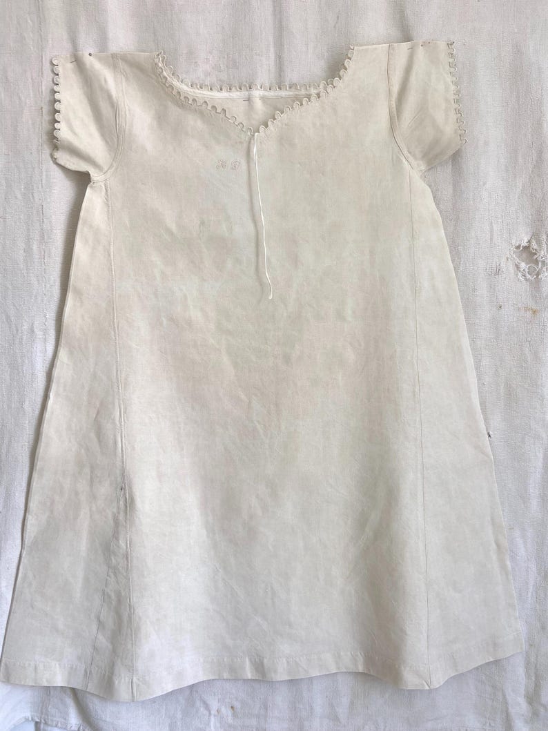 Puede incluir: Un vestido recto de color blanco roto con mangas cortas, que presenta un escote festoneado con un borde delicado. La prenda tiene una silueta sencilla y recta con costuras verticales en la parte delantera y un fino lazo en el centro del cuello.