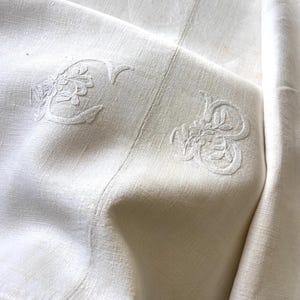 May include: Close-up of a white linen textile with embroidered floral monograms. The fabric is folded, showing texture and detail of the stitching. The embroidery is in a matching white thread.