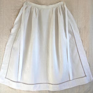 Op de afbeelding: Een witte schort met een rechthoekige vorm en een decoratieve gestikte rand. De schort heeft lange, dunne banden en is gemaakt van een lichte stof. De schort is waarschijnlijk vintage of handgemaakt.