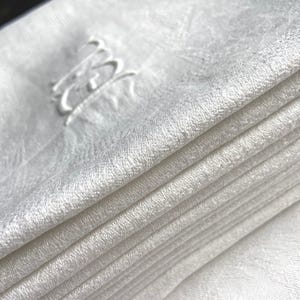 May include: A stack of folded white linen napkins. Each napkin has a subtle, textured pattern and is embroidered with a delicate, cursive monogram in the upper corner. The napkins are neatly stacked, showcasing their clean, crisp appearance.
