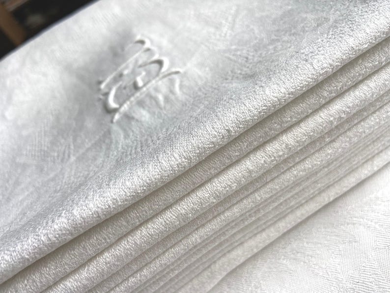 May include: A stack of folded white linen napkins. Each napkin has a subtle, textured pattern and is embroidered with a delicate, cursive monogram in the upper corner. The napkins are neatly stacked, showcasing their clean, crisp appearance.