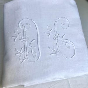 Può includere: Federa in lino bianco con iniziali "A" e "S" ricamate. Le iniziali sono in una scrittura decorativa con accenti floreali e di foglie. La federa è piegata, mostrando il ricamo.