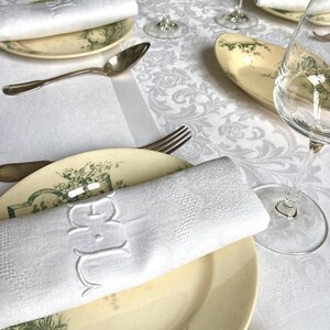 May include: Elegant table setting with white monogrammed napkins, antique plates with green botanical designs, silverware, and wine glasses. The tablecloth features a subtle floral pattern, creating a refined dining atmosphere.
