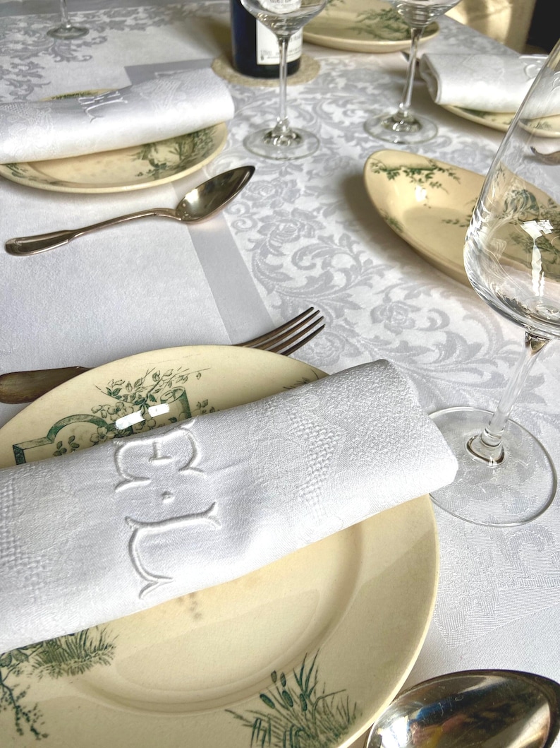 May include: Elegant table setting with white monogrammed napkins, antique plates with green botanical designs, silverware, and wine glasses. The tablecloth features a subtle floral pattern, creating a refined dining atmosphere.