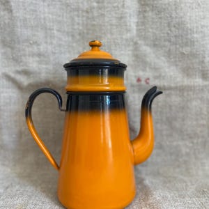 Puede incluir: Cafetera de esmalte naranja y negro con tapa. La cafetera tiene un cuerpo redondeado y un pico. El asa está unida al lado de la cafetera.
