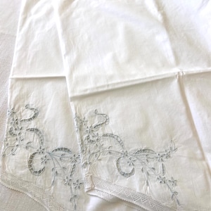 May include: Two white pillowcases with delicate blue floral embroidery and lace trim along the bottom edge. The pillowcases are folded and layered, showcasing the intricate details of the design.