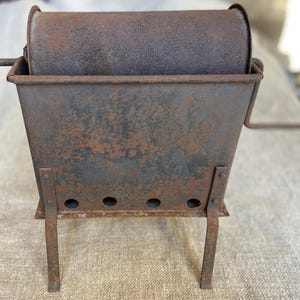 Op de afbeelding: Een vintage, roestige metalen koffierooster met een handcrank. De roaster heeft een cilindrische trommel en een rechthoekige basis met vier poten.
