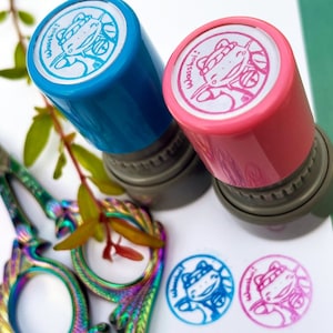 May include: Two colorful ink stamps with a cartoon frog design. The stamps are blue and pink with the text "Wasshoi!" on each. The stamps are on a white surface with two additional frog stamps in blue and pink.