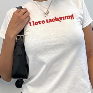 Op de afbeelding: Een wit T-shirt met korte mouwen en de tekst i love taehyung in het rood op de borst gedrukt. Het shirt heeft een ronde hals en een standaard pasvorm. Een zwarte schoudertas met een zilveren gesp bevindt zich aan de zijkant van het kledingstuk.