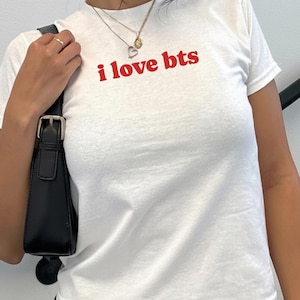 May include: A white short-sleeved t-shirt featuring the text i love bts printed in red on the chest. The shirt has a crew neckline and is paired with a black shoulder bag with a silver buckle detail and layered gold-toned necklaces.