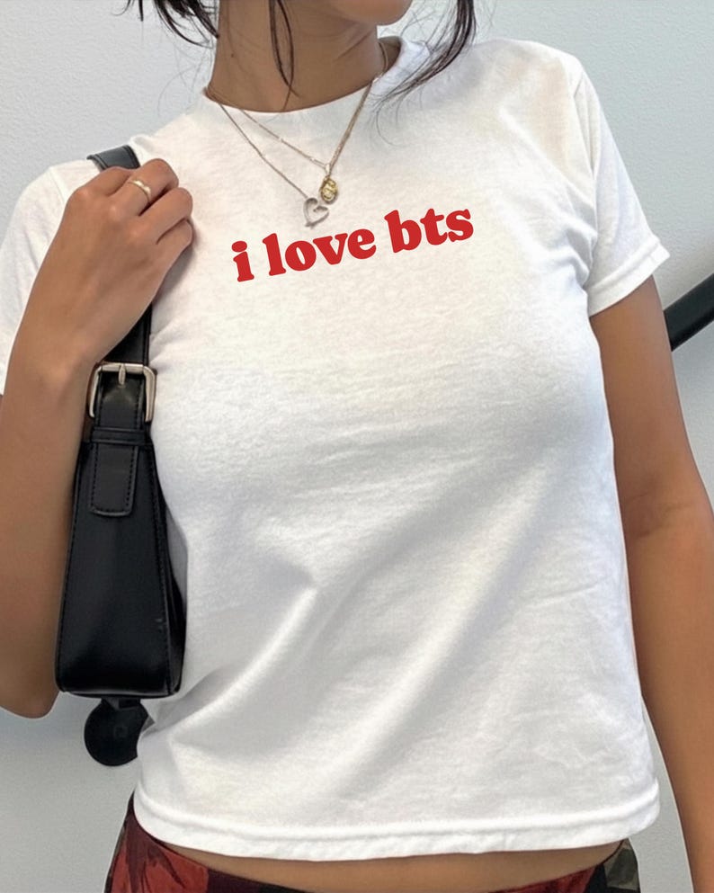 May include: A white short-sleeved t-shirt featuring the text i love bts printed in red on the chest. The shirt has a crew neckline and is paired with a black shoulder bag with a silver buckle detail and layered gold-toned necklaces.