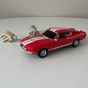 May include: A red toy car with white racing stripes, a black windscreen, and black wheels. The car has a cord and a three-pin plug attached. The car is a classic model.