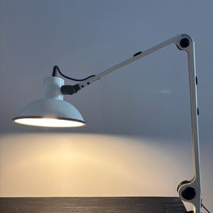 Può includere: Una lampada da scrivania bianca con braccio articolato regolabile e paralume a cupola. La lampada presenta giunti meccanici neri e una base a morsetto per il fissaggio a una superficie. La luce è accesa e proietta un bagliore caldo.