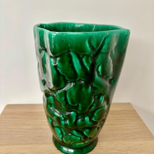 Könnte beinhalten: Eine glänzende, smaragdgrüne Keramikvase mit einem strukturierten, organischen Design. Die Vase hat eine breite Öffnung und einen Sockel. Die Oberfläche hat ein erhabenes, blattartiges Muster. Die Vase wird auf einer hellbraunen Holzoberfläche ausgestellt.
