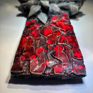 May include: A ceramic wall sculpture with a textured, red and black surface. The sculpture features a raised, abstract design with a three-dimensional effect.