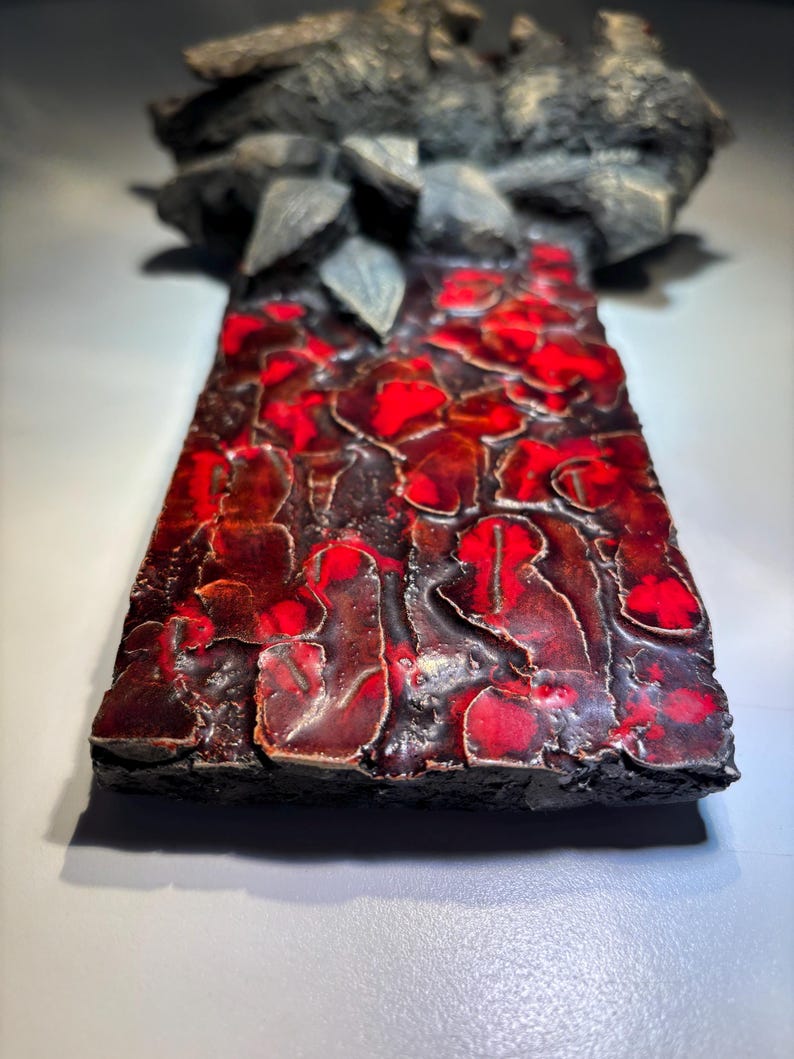May include: A ceramic wall sculpture with a textured, red and black surface. The sculpture features a raised, abstract design with a three-dimensional effect.