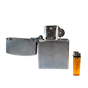 Op de afbeelding: Een zilveren aansteker met een geborstelde afwerking en een apart deksel. Ernaast ligt een kleine oranje aansteker.