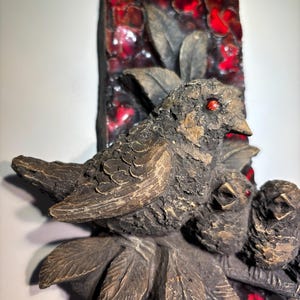 May include: A ceramic wall sculpture of two birds perched on a branch with red and brown leaves. The birds are brown with red eyes and are sculpted in a realistic style.