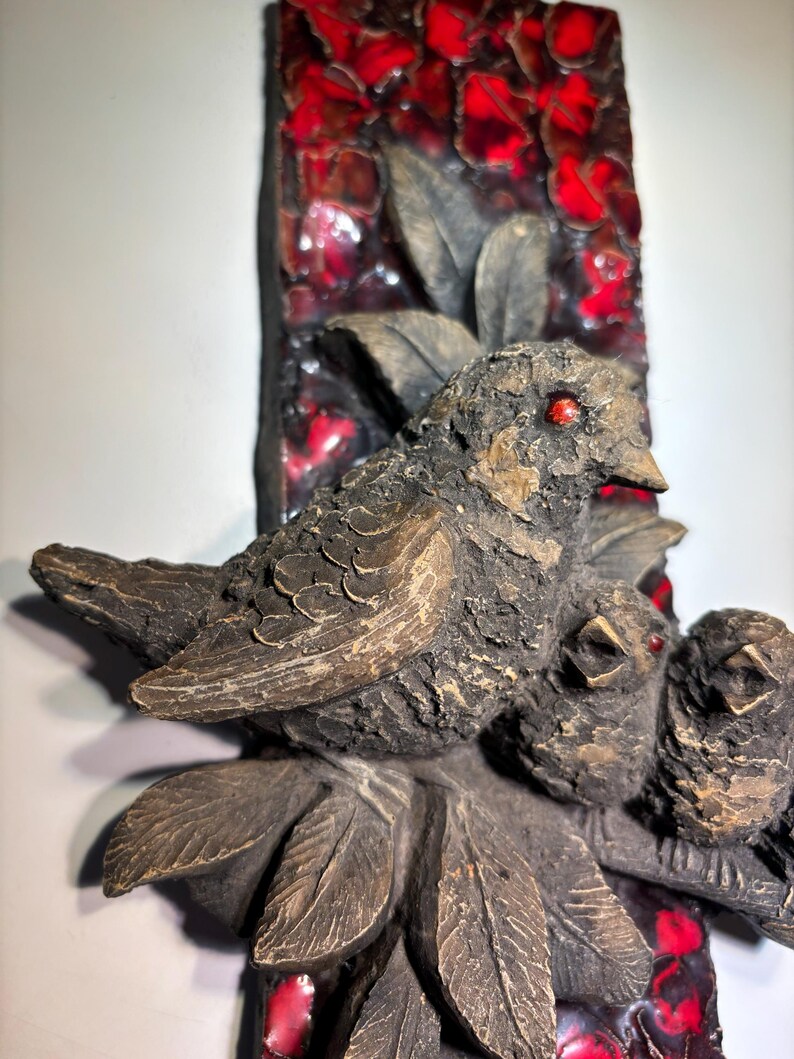 May include: A ceramic wall sculpture of two birds perched on a branch with red and brown leaves. The birds are brown with red eyes and are sculpted in a realistic style.
