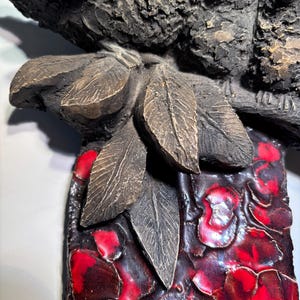 May include: A ceramic wall sculpture featuring two birds perched on a branch with leaves. The birds are brown and have red accents. The background is a textured red and black design.