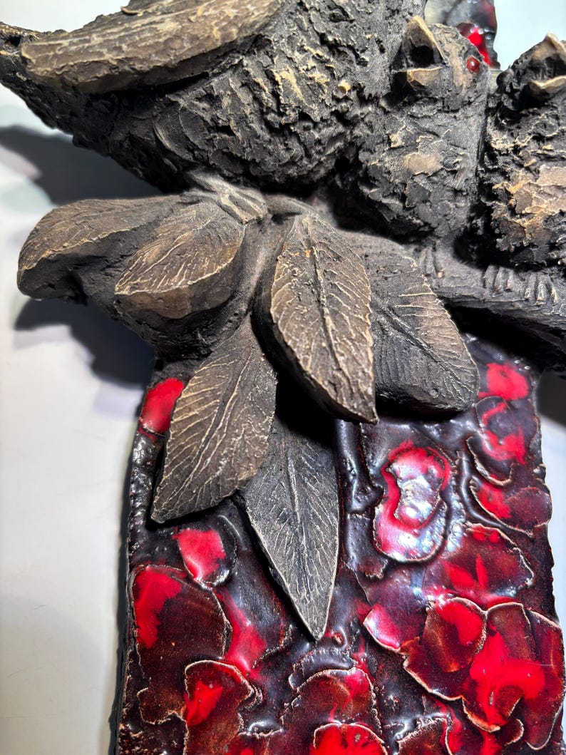 May include: A ceramic wall sculpture featuring two birds perched on a branch with leaves. The birds are brown and have red accents. The background is a textured red and black design.