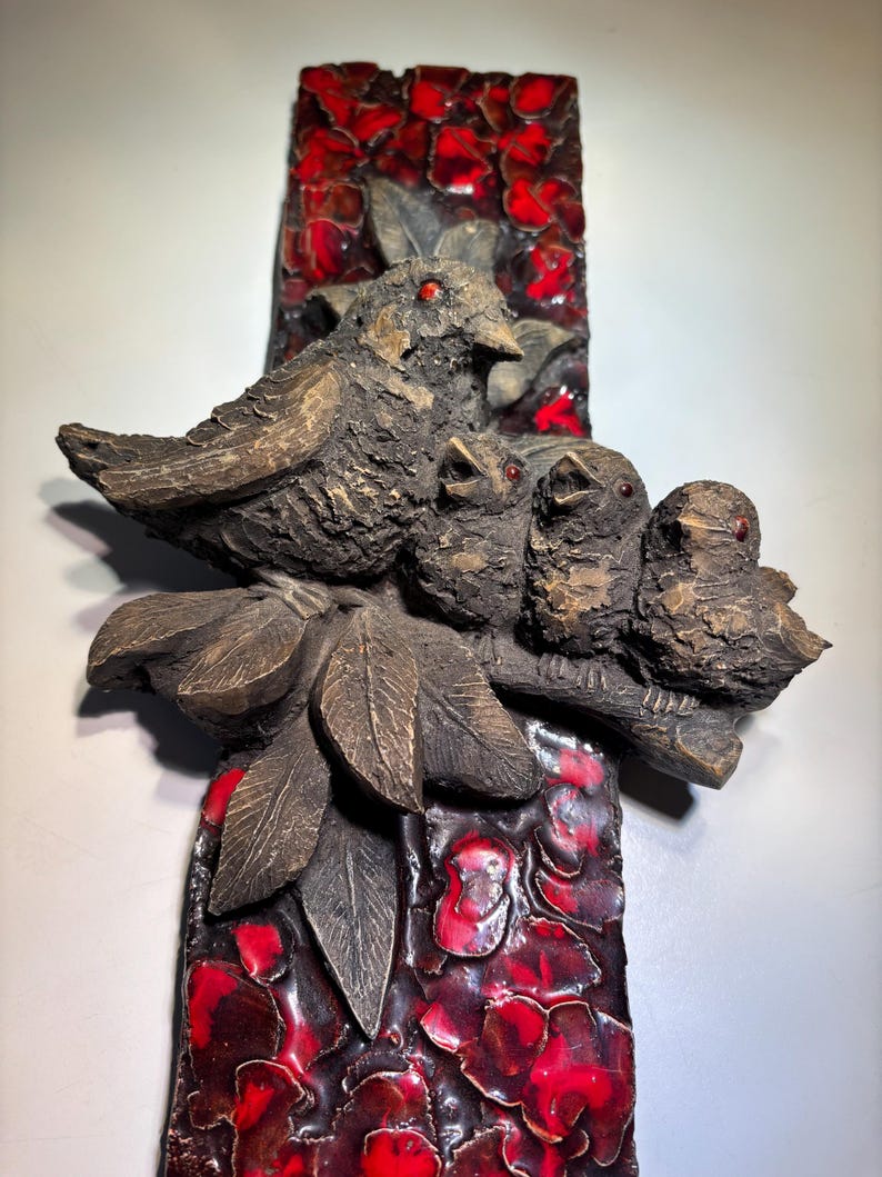 May include: A ceramic wall sculpture featuring a family of birds perched on a branch with red floral background.