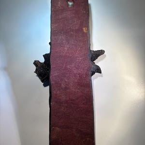 May include: A dark brown wooden wall hanging with a textured surface. The hanging has a burgundy backing and a metal hook on the top.