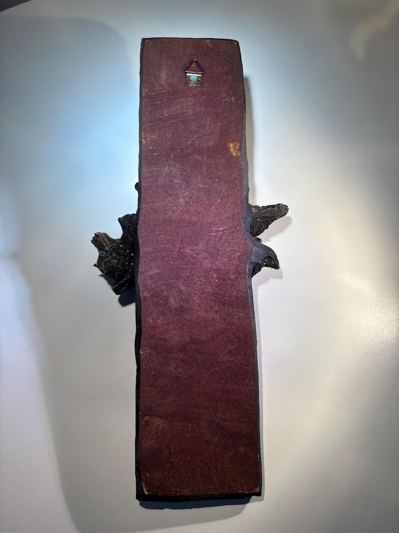 May include: A dark brown wooden wall hanging with a textured surface. The hanging has a burgundy backing and a metal hook on the top.