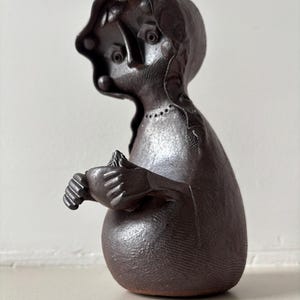 Könnte beinhalten: Eine dunkelbraune Keramikskulptur einer stilisierten menschlichen Figur. Die Figur hat einen abgerundeten Körper, einen großen Kopf mit Gesichtszügen und Arme, die ein kleines Objekt halten. Die Skulptur hat eine strukturierte Oberfläche.