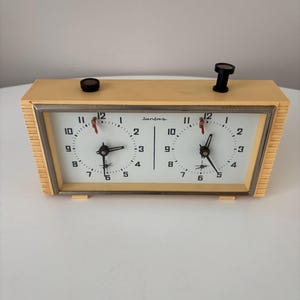 Op de afbeelding: Een vintage schaakklok met een crèmekleurige behuizing en twee witte wijzerplaten. De klok heeft zwarte knoppen aan de bovenkant en een zilveren frame rond de wijzerplaten. Het woord "Jantar" staat op de wijzerplaat.