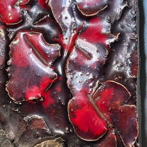 May include: Close-up of a ceramic tile with a textured surface. The tile is decorated with a red and black glaze, creating a abstract pattern.