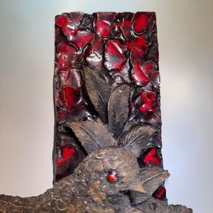 May include: A ceramic wall sculpture featuring a group of birds perched on a branch. The background is a red and black mosaic tile design.