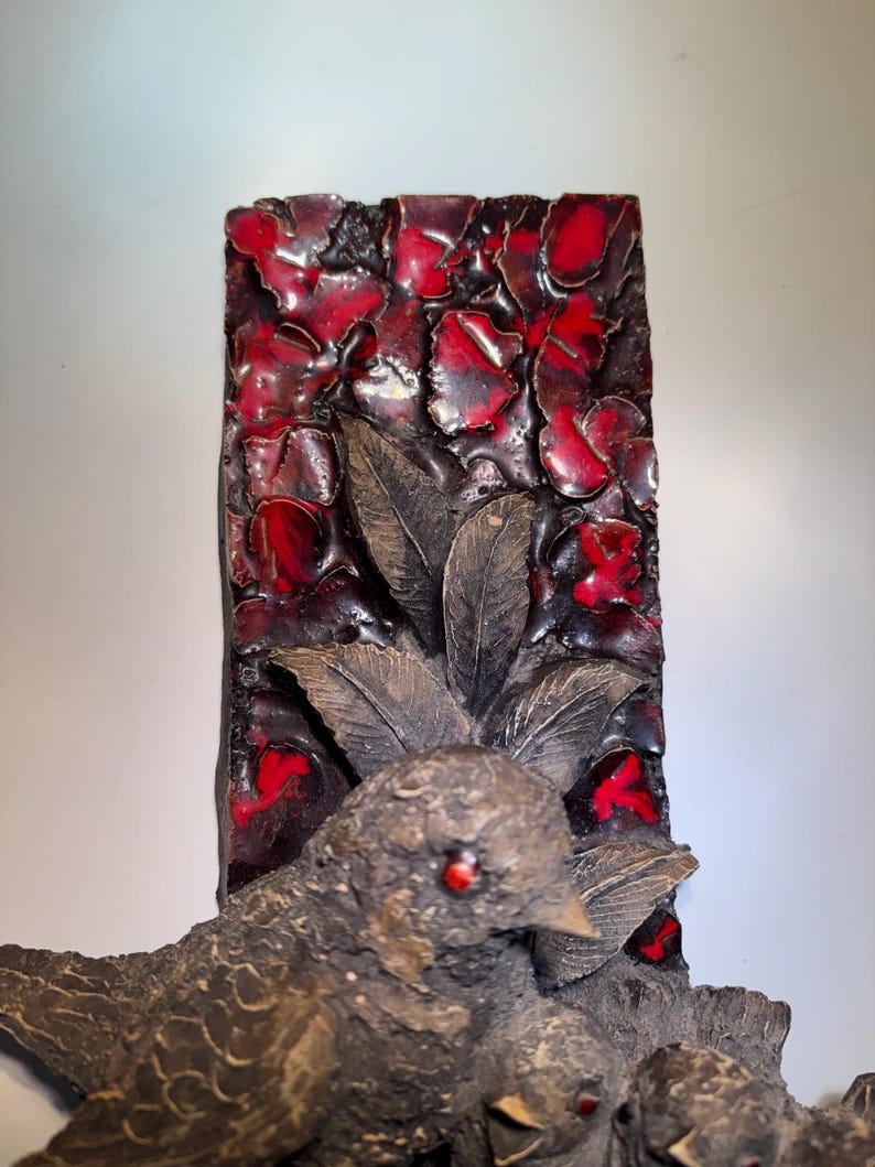 May include: A ceramic wall sculpture featuring a group of birds perched on a branch. The background is a red and black mosaic tile design.
