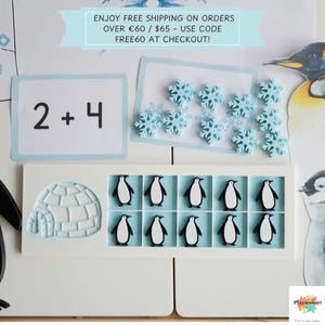 Op de afbeelding: Educatief speelgoedset met een wit frame met pinguïnfiguren, een iglo en een wiskundige opgave. Lichtblauwe sneeuwvlokvormige tellers en een pinguïnillustratie zijn ook aanwezig. De tekst "2 + 4" is zichtbaar.