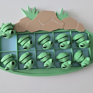 Pode incluir: Um brinquedo com tema de sapo verde com uma base oval verde claro e uma grade de dez quadrados. Cada quadrado contém um sapo verde claro. Acima da grade há uma formação rochosa marrom com detalhes em grama verde. O brinquedo é para crianças.