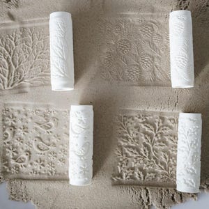 May include: White ceramic rolling pins with embossed nature-themed designs. The designs include branches, pine cones, birds, snowflakes, and holly leaves. The rolling pins are displayed on a textured, light brown surface.