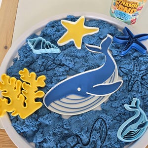 Puede incluir: Un juego sensorial con arena cinética azul y moldes con temática oceánica, incluyendo una ballena, estrellas de mar y coral. Un recipiente de arena dinámica azul es visible. El conjunto está dispuesto sobre una bandeja blanca.