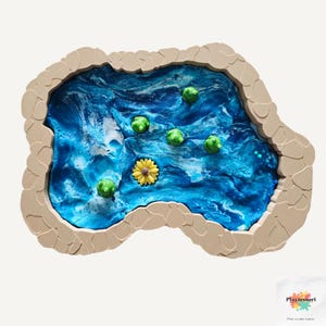 Puede incluir: Un juguete sensorial artesanal que presenta un estanque de resina azul con nenúfares verdes y una flor amarilla. El estanque está enmarcado por un borde beige con forma de piedra. El juguete está diseñado para el juego y el aprendizaje de los niños.