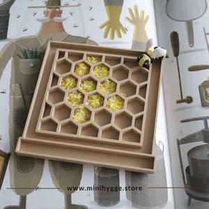 Puede incluir: Una bandeja de panal de abeja de silicona beige con una pequeña figura de abeja amarilla y negra en la parte superior. La bandeja está llena de arroz blanco sin cocinar. La bandeja está en una página de un libro infantil con ilustraciones de abejas y otros insectos. El texto en la página dice "www.minihygge.store".