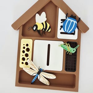 Op de afbeelding: Een bruin, huisvormig insectenhotel met een geel honingraatpaneel, een wit centraal paneel met een verticale gleuf, een zwart rooster en kleurrijke insectenfiguren, waaronder een bij, een blauwe kever, een groene sprinkhaan en een libel.