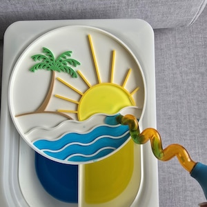Op de afbeelding: Een witte, rechthoekige tray met een cirkelvormig, gelaagd ontwerp dat een strandscène weergeeft met een palmboom, zon en golven. De tray heeft ook een blauw en geel gedeelte en een gebogen, oranje en groene buis.