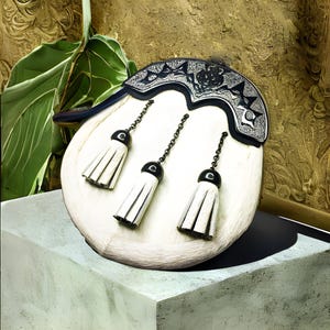 May include: A cream-colored sporran with a decorative silver and black cantle. The sporran features three white tassels with black caps and chain attachments. The background includes a green plant and a textured gold surface.