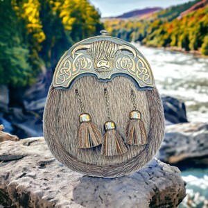 Puede incluir: Un sporran de kilt escocés tradicional con una parte superior de metal plateado que presenta un diseño de vaca de las Tierras Altas y nudos celtas. El sporran tiene un cuerpo de piel marrón y tres borlas decorativas.