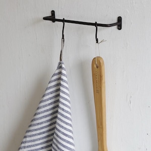 May include: A black metal wall-mounted towel rack with two hooks. A blue and white striped towel hangs from one hook. A wooden handled body brush with natural bristles hangs from the other hook.